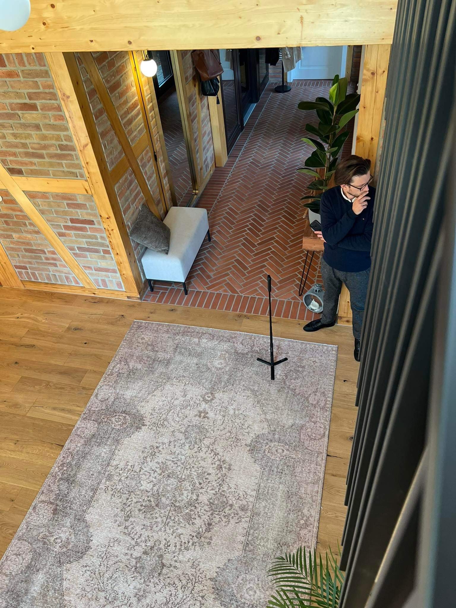 Wnętrze domu z widokiem z góry: parkiet, dywan w stylu vintage, ceglana ściana z drewnianą konstrukcją, mężczyzna w okularach oparty o balustradę.