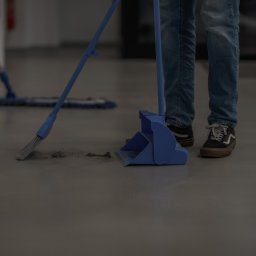 HUB2WORK SPÓŁKA Z OGRANICZONĄ ODPOWIEDZIALNOŚCIĄ - Sprzątanie podłogi w biurze: osoba zamiata szarą podłogę niebieską zmiotką i szufelką. W tle widoczny mop. Ujęcie od dołu, skupione na narzędziach i etapie pracy.