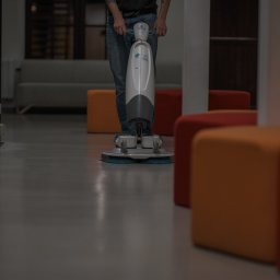 HUB2WORK SPÓŁKA Z OGRANICZONĄ ODPOWIEDZIALNOŚCIĄ - Pracownik sprząta podłogę w biurze za pomocą automatycznej maszyny czyszczącej. W tle widoczne schody i kolorowe pufy. Szara, lśniąca podłoga.