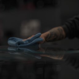 HUB2WORK SPÓŁKA Z OGRANICZONĄ ODPOWIEDZIALNOŚCIĄ - Tatuowana dłoń czyści błyszczącą, ciemną powierzchnię niebieską ściereczką z mikrofibry. Delikatne światło podkreśla czystość.