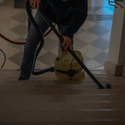 HUB2WORK SPÓŁKA Z OGRANICZONĄ ODPOWIEDZIALNOŚCIĄ - Osoba odkurza schody odkurzaczem przemysłowym w budynku w Warszawie. W tle widoczny czerwony hydrant i marmurowe schody.