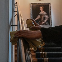 HUB2WORK SPÓŁKA Z OGRANICZONĄ ODPOWIEDZIALNOŚCIĄ - Pracownik w trakcie czyszczenia poręczy schodów w budynku. W tle obraz w ramie. Osoba ma identyfikator ze swoim imieniem. Używa żółtej ściereczki i rozpyla preparat.