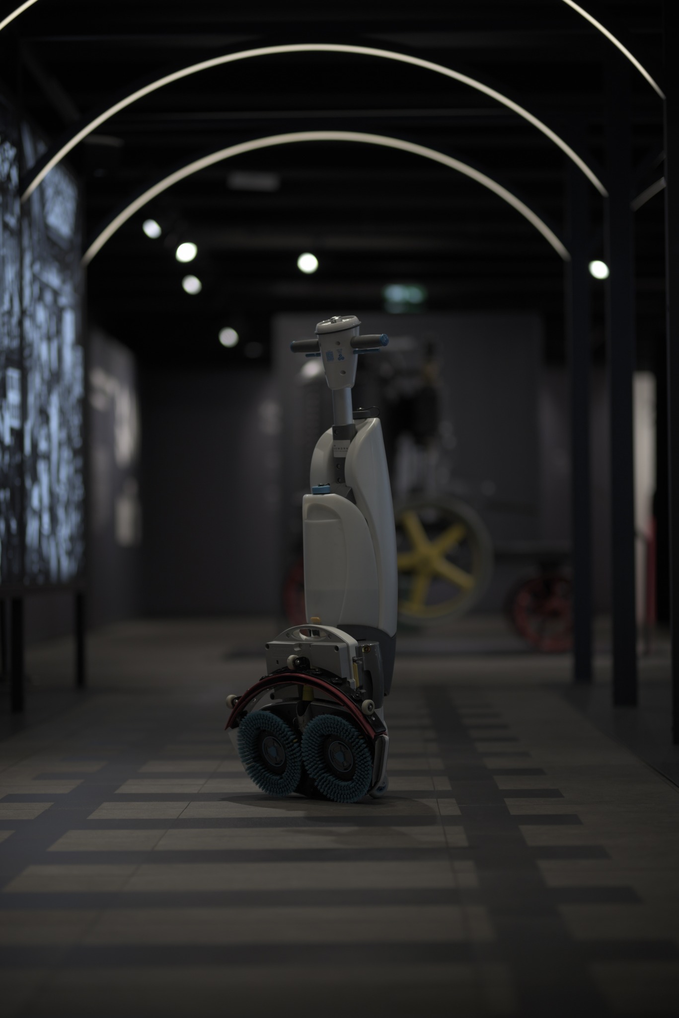 Profesjonalna maszyna do czyszczenia podłóg z dwiema okrągłymi szczotkami na tle korytarza z oświetleniem łukowym. Widoczne detale konstrukcyjne i elementy sterujące.