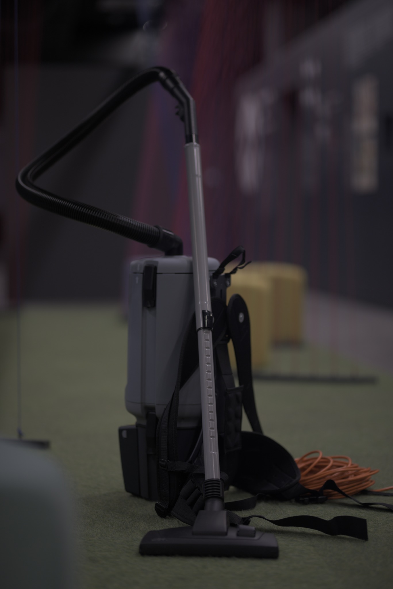 Profesjonalny odkurzacz plecakowy z kablem zasilającym na zielonej wykładzinie, gotowy do użycia w pomieszczeniu biurowym lub innym obiekcie komercyjnym.