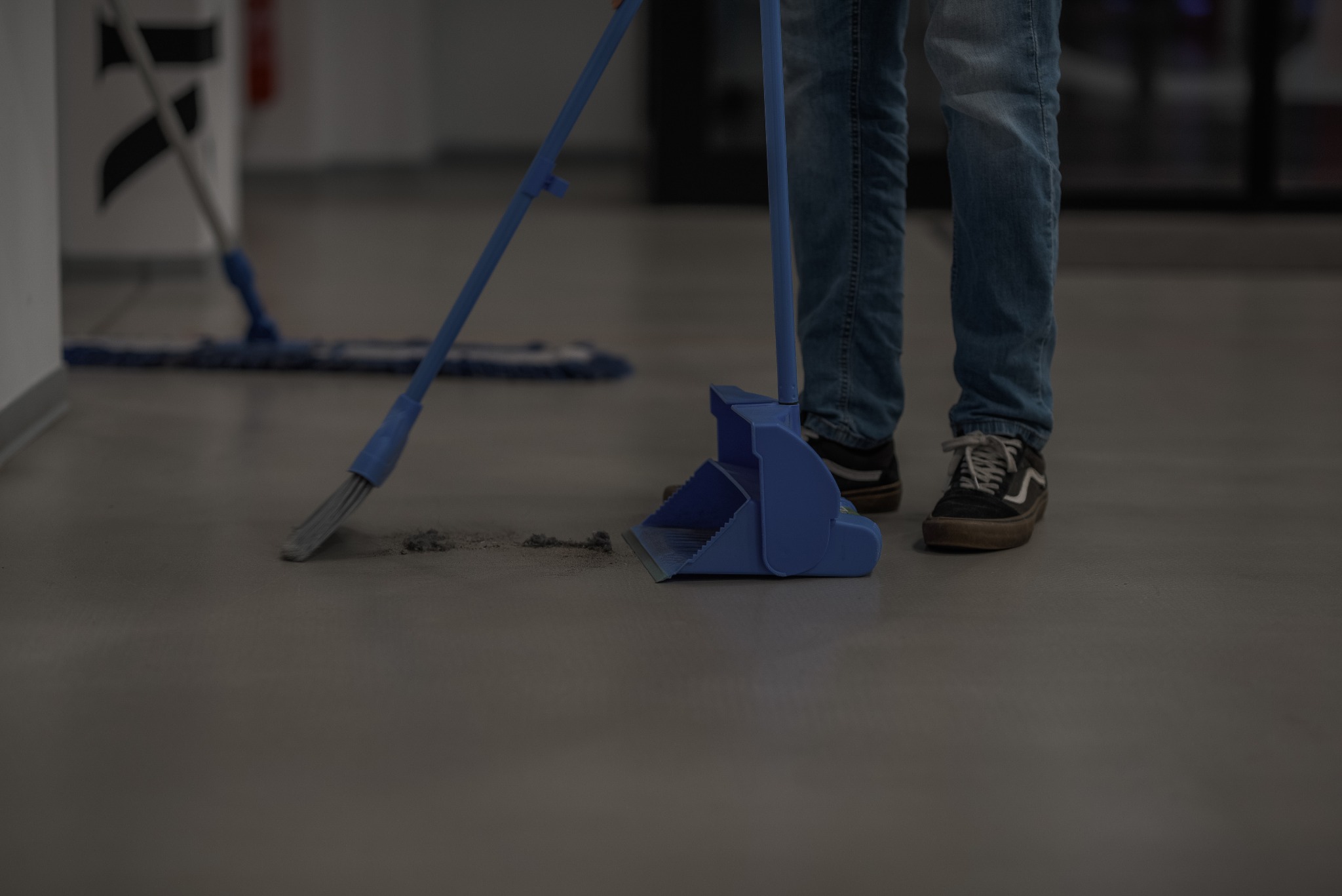 Sprzątanie podłogi w biurze: osoba zamiata szarą podłogę niebieską zmiotką i szufelką. W tle widoczny mop. Ujęcie od dołu, skupione na narzędziach i etapie pracy.