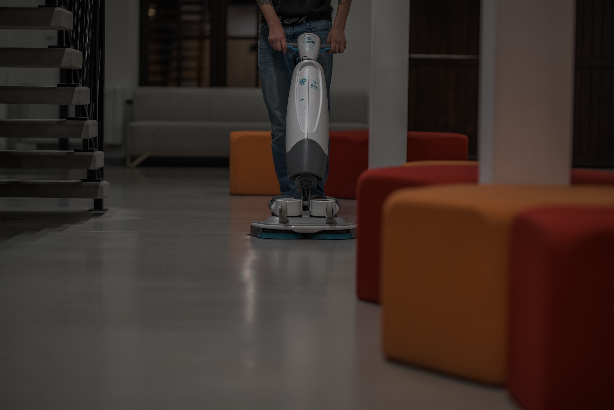 Pracownik sprząta podłogę w biurze za pomocą automatycznej maszyny czyszczącej. W tle widoczne schody i kolorowe pufy. Szara, lśniąca podłoga.