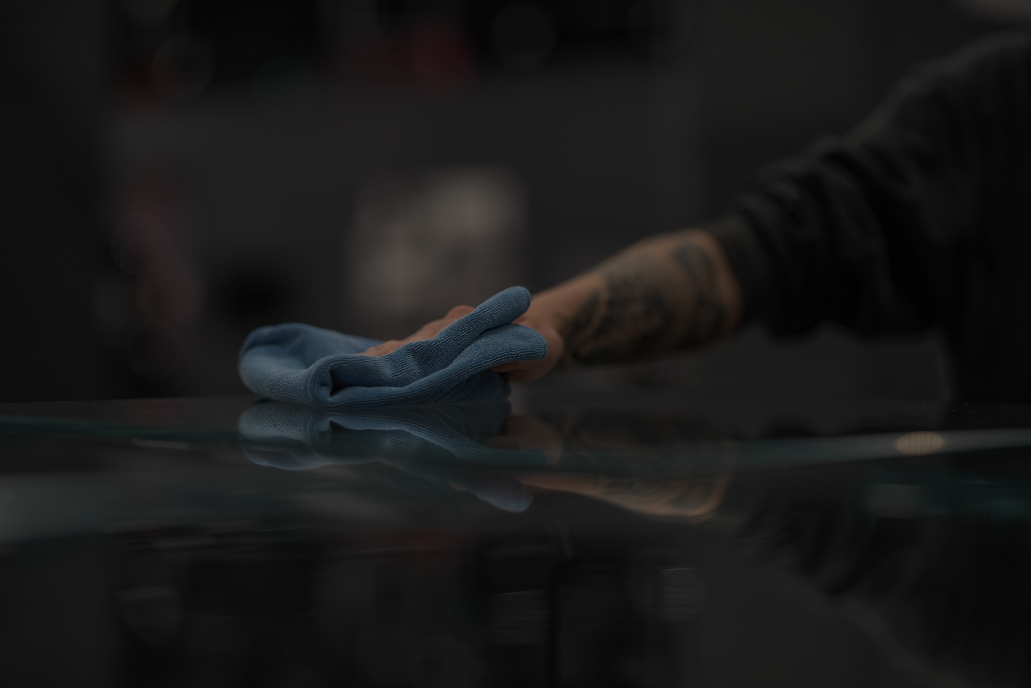 Tatuowana dłoń czyści błyszczącą, ciemną powierzchnię niebieską ściereczką z mikrofibry. Delikatne światło podkreśla czystość.