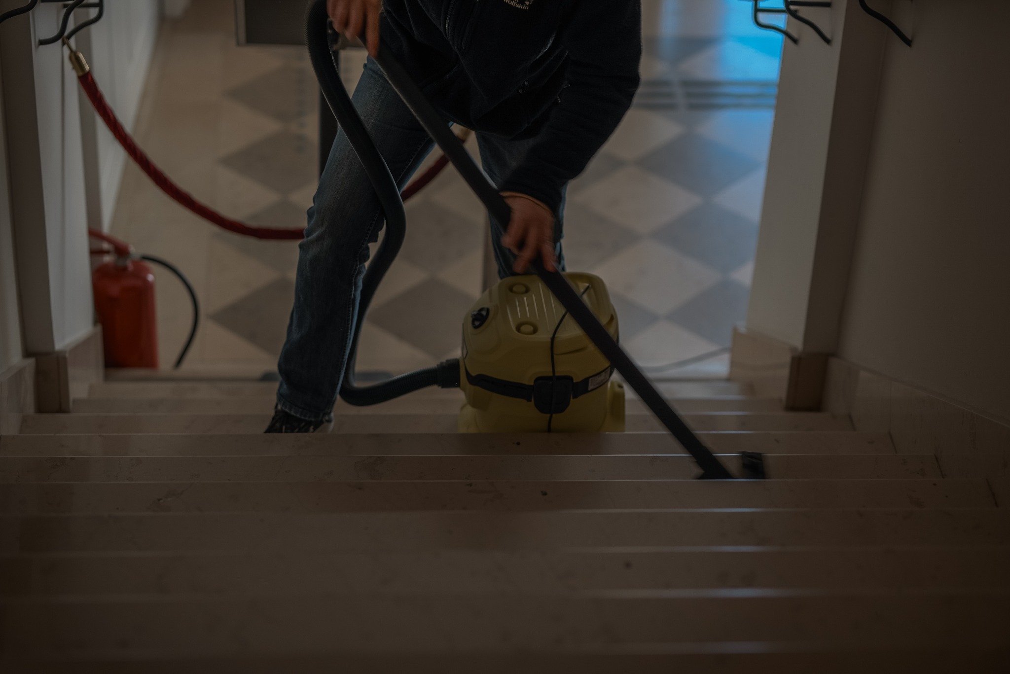 Osoba odkurza schody odkurzaczem przemysłowym w budynku w Warszawie. W tle widoczny czerwony hydrant i marmurowe schody.