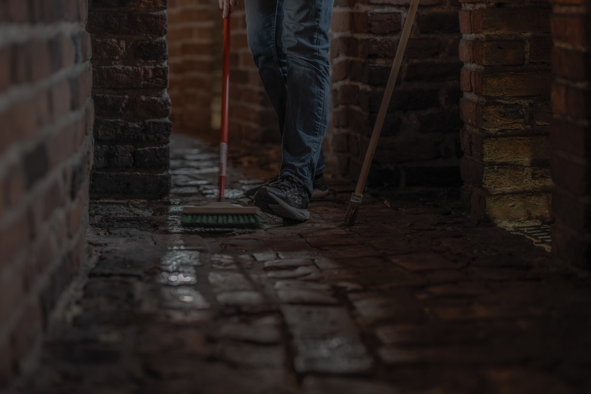 Czyszczenie ceglanej podłogi w przejściu. Widoczne narzędzia: szczotka z czerwonym trzonkiem i miotła. Osoba w jeansach i sportowych butach. Ciemne, klimatyczne wnętrze.