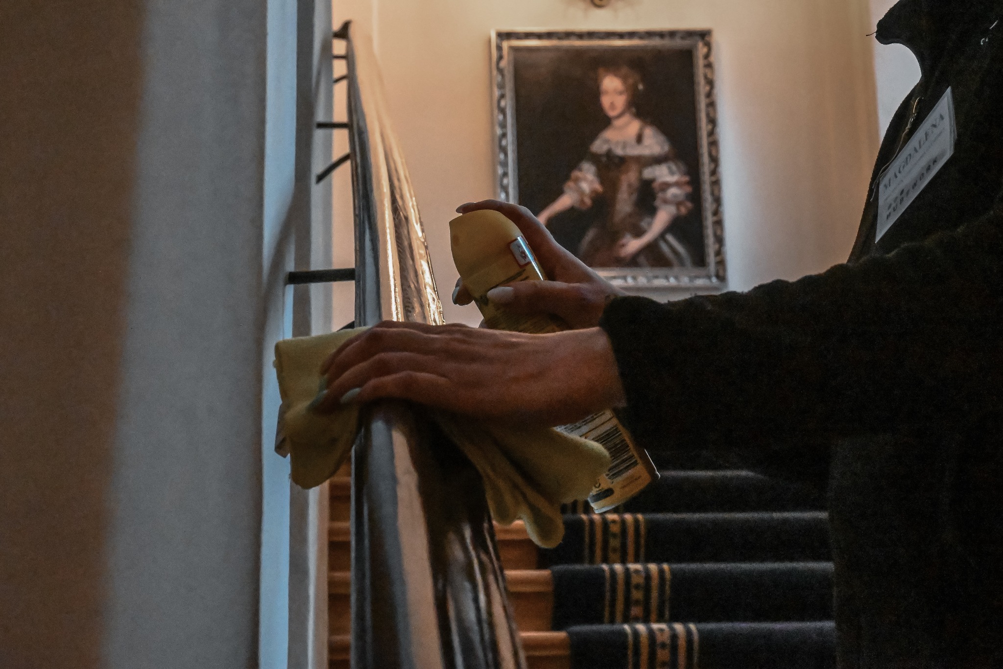 Pracownik w trakcie czyszczenia poręczy schodów w budynku. W tle obraz w ramie. Osoba ma identyfikator ze swoim imieniem. Używa żółtej ściereczki i rozpyla preparat.