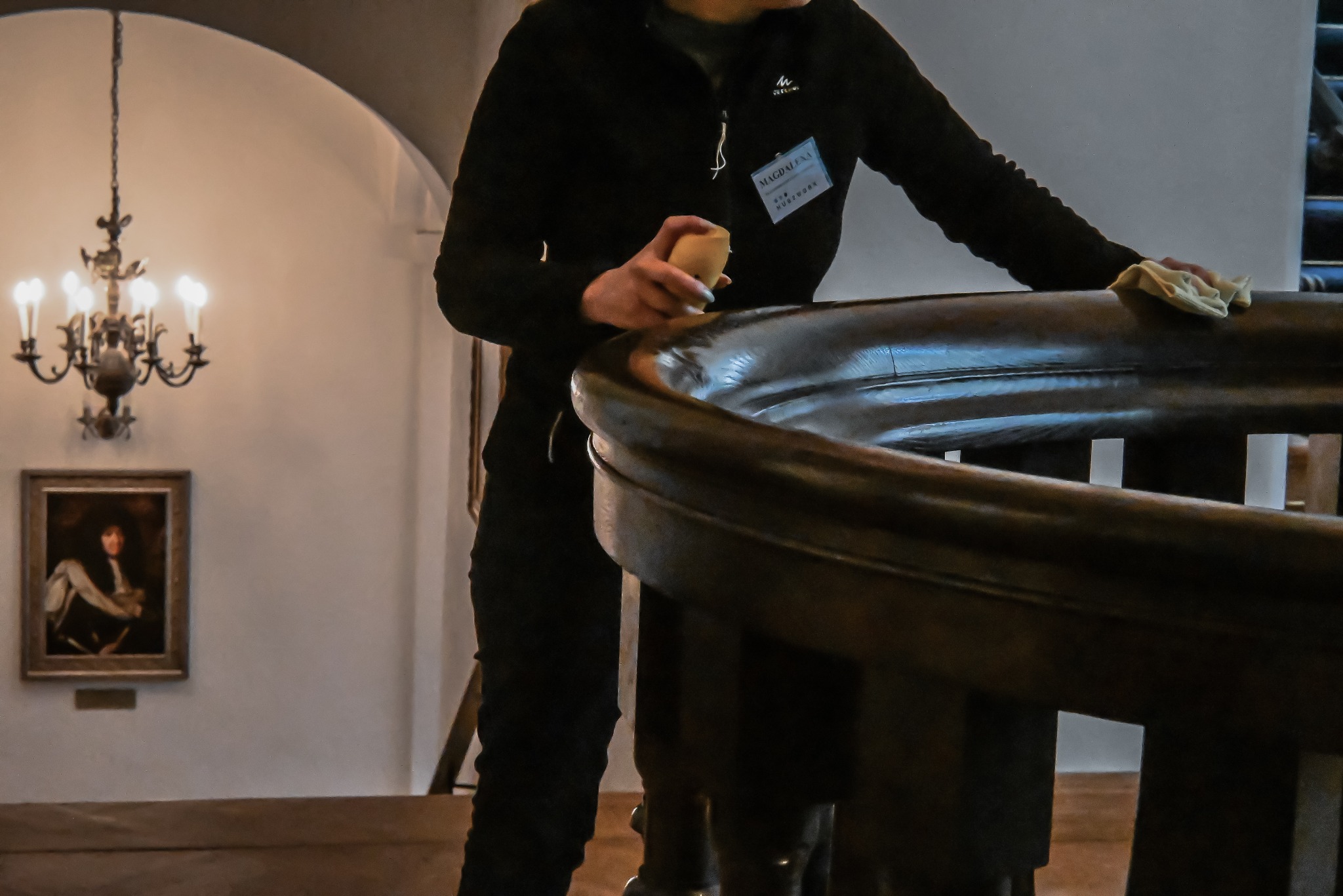 Pracownik w trakcie czyszczenia drewnianej balustrady schodów w budynku z obrazami i żyrandolem. Osoba ma identyfikator. Używa ścierki i preparatu.