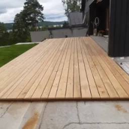 Nowo ułożony taras z jasnego drewna z widoczną siatką zbrojeniową i regulowanymi wspornikami pod konstrukcją, na tle domu z czarną elewacją i zielenią.