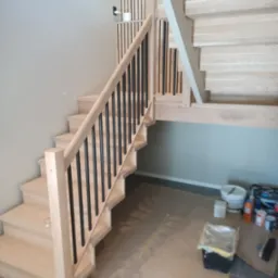 Jasne drewniane schody z czarnymi metalowymi balustradami w trakcie montażu, widoczne zabezpieczenia podłogi i akcesoria malarskie.