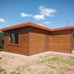 Andrzej Miśkiewicz - Gotowy, drewniany domek z brązowymi elementami wykończeniowymi, widoczny na trawniku w słoneczny dzień. Obok huśtawka i drabina. Widoczne okno i drzwi.