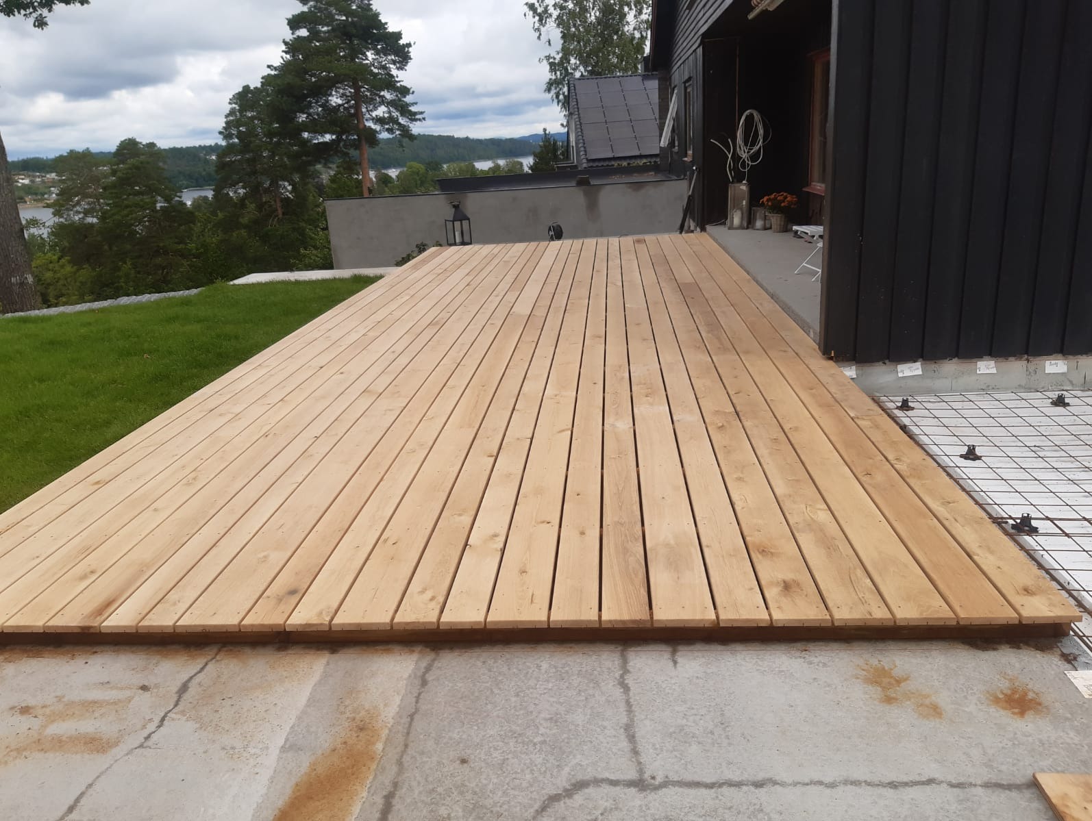 Nowo ułożony taras z jasnego drewna z widoczną siatką zbrojeniową i regulowanymi wspornikami pod konstrukcją, na tle domu z czarną elewacją i zielenią.