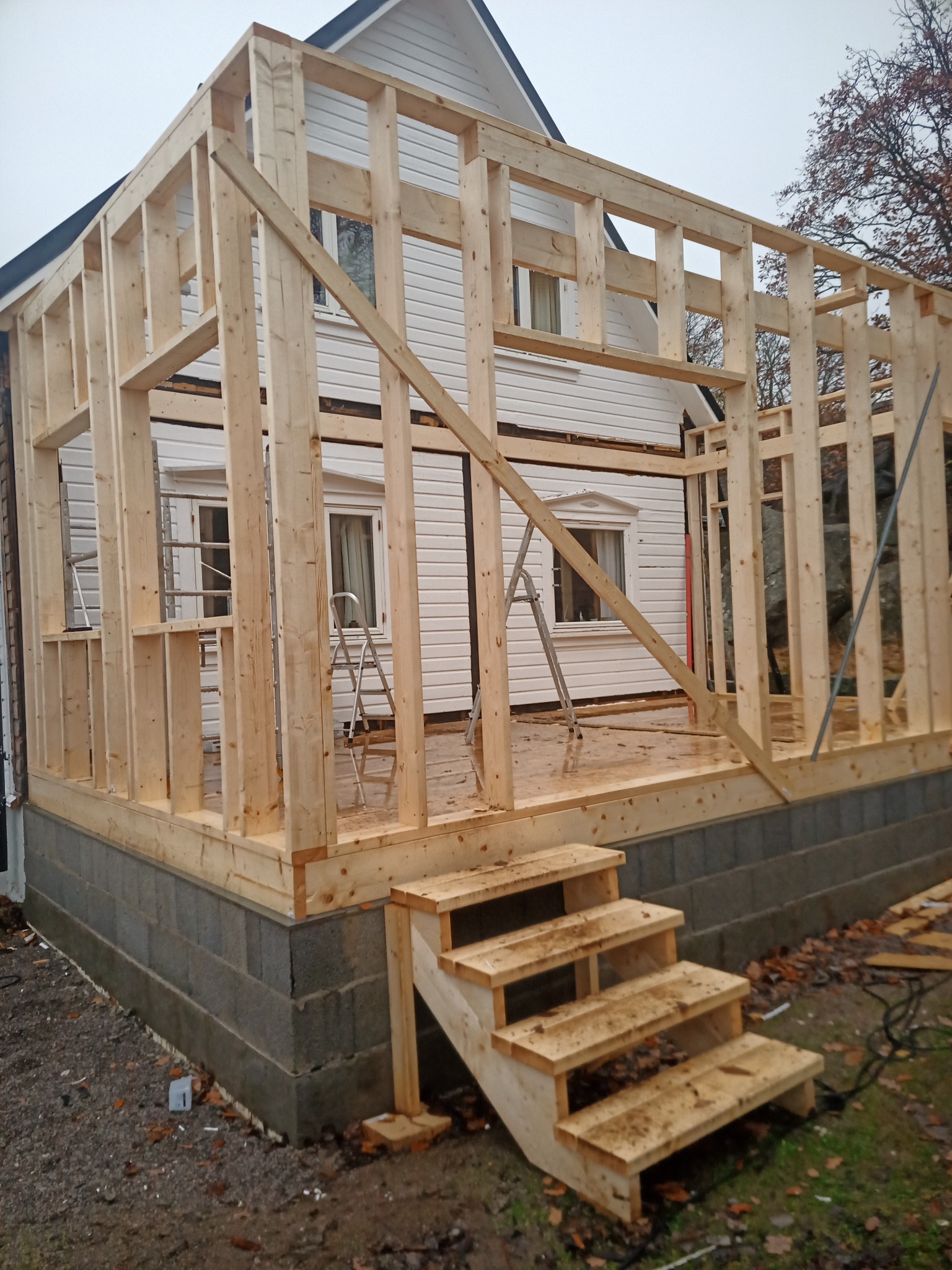 Surowa konstrukcja szkieletowa domu z widocznymi schodami wejściowymi, oparta na fundamencie z szarych bloczków betonowych, z istniejącym domem w tle i dwiema aluminiowymi drabinami wewnątrz budowy.