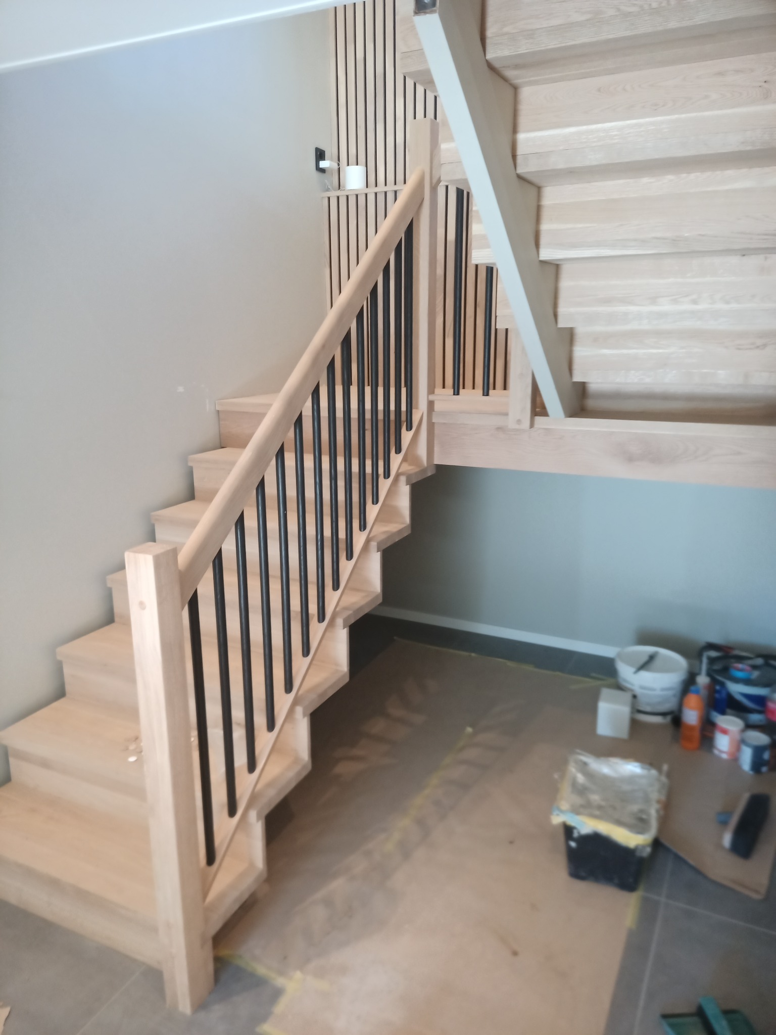 Jasne drewniane schody z czarnymi metalowymi balustradami w trakcie montażu, widoczne zabezpieczenia podłogi i akcesoria malarskie.