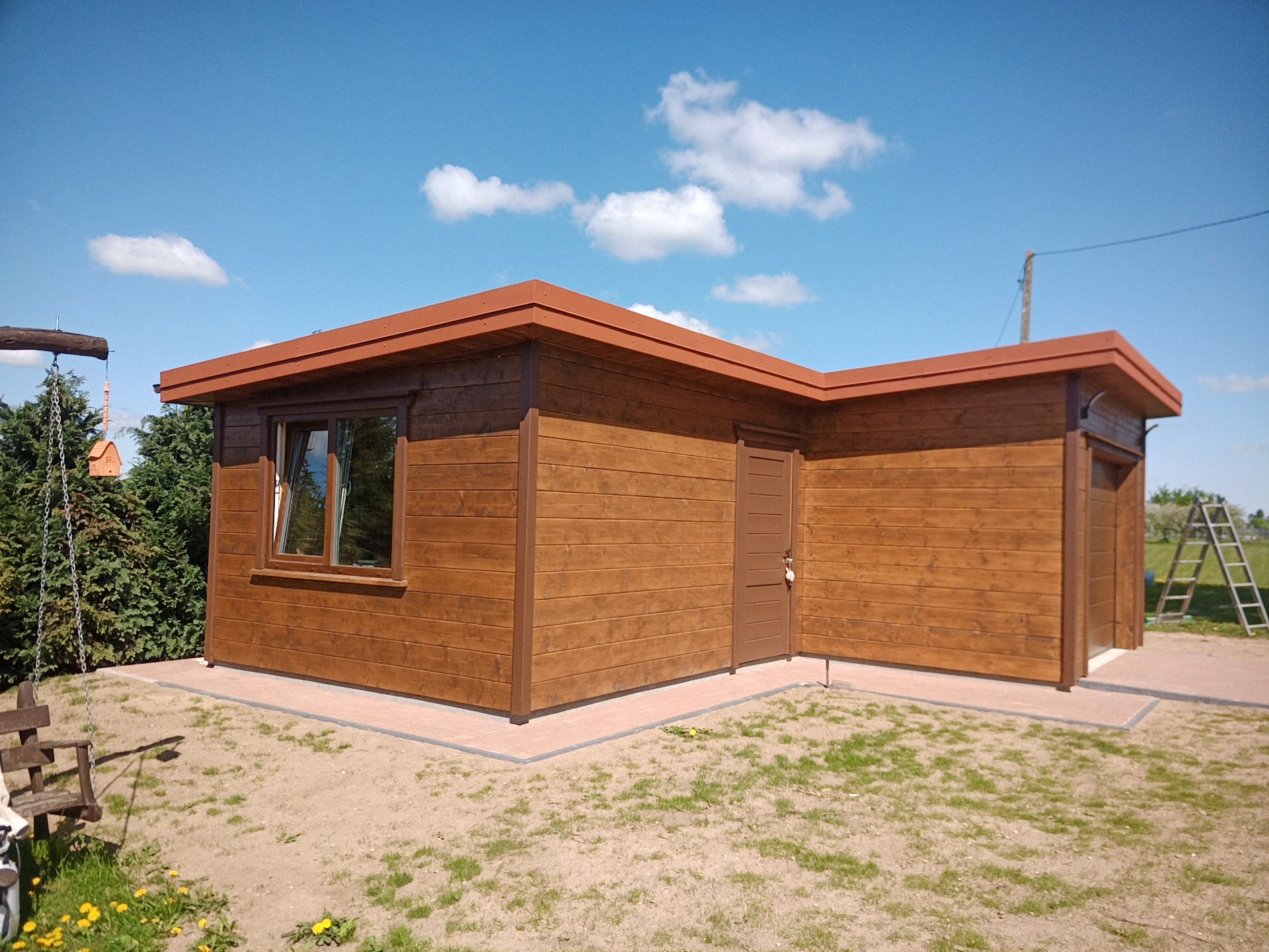 Gotowy, drewniany domek z brązowymi elementami wykończeniowymi, widoczny na trawniku w słoneczny dzień. Obok huśtawka i drabina. Widoczne okno i drzwi.