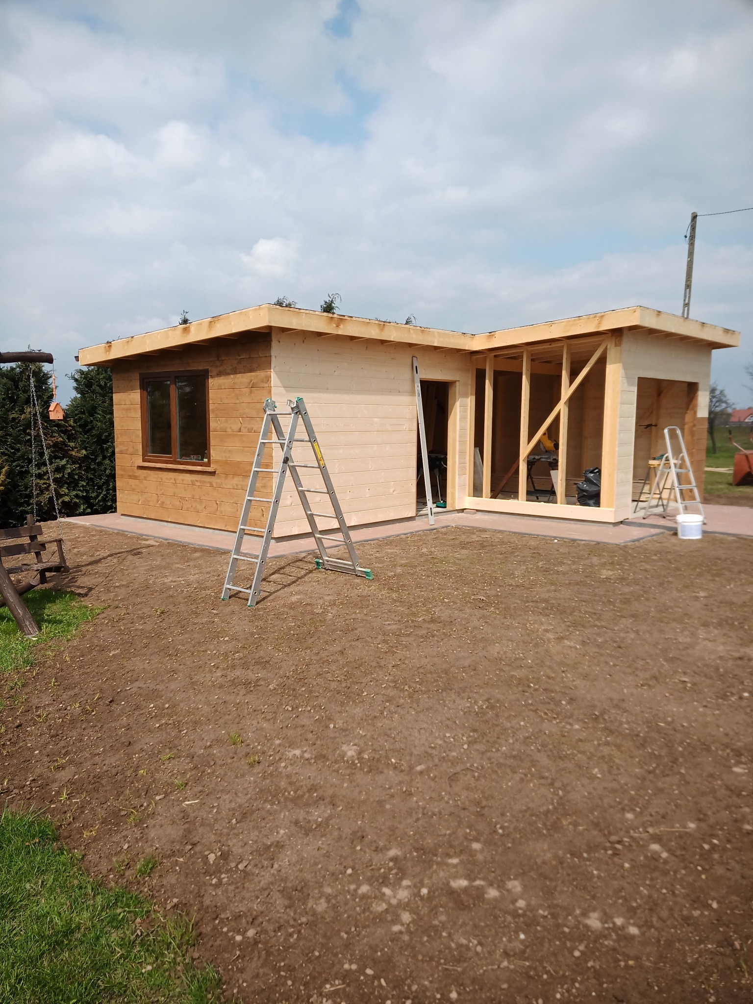 Surowa konstrukcja drewniana budynku w kształcie litery L, częściowo pomalowana bejcą, z widocznymi schodami i drabinkami, otoczona przygotowanym terenem pod trawnik.