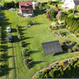Mariusz Oraczewski - Widok z lotu ptaka na duży ogród z panelem słonecznym, dwoma samochodami na podjeździe i domami w tle.