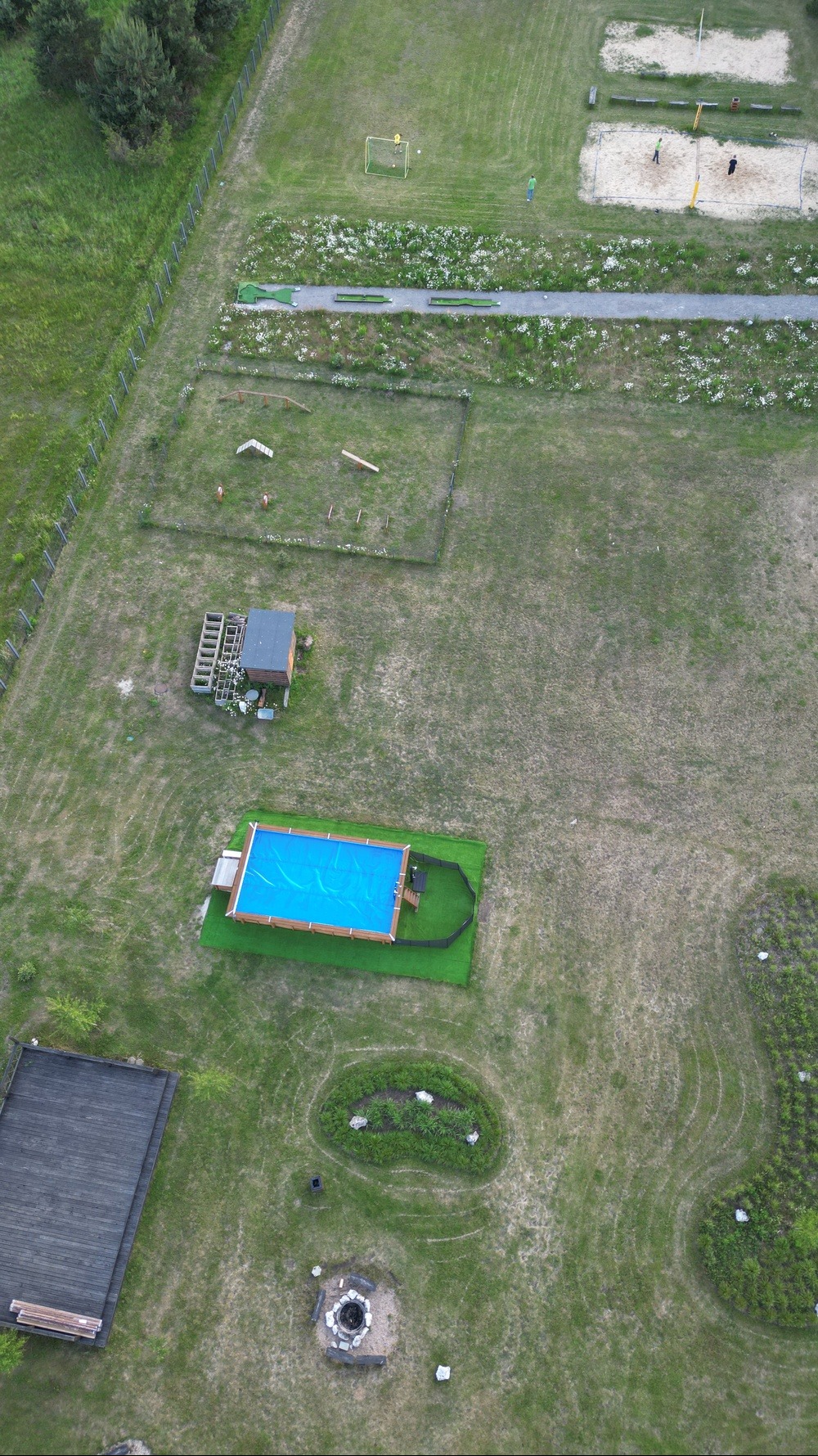 Widok z lotu ptaka na duży ogród z basenem z niebieską wodą, placem zabaw dla dzieci z piaskownicą i boiskiem do piłki nożnej, miejscem na ognisko i drewnianym tarasem.