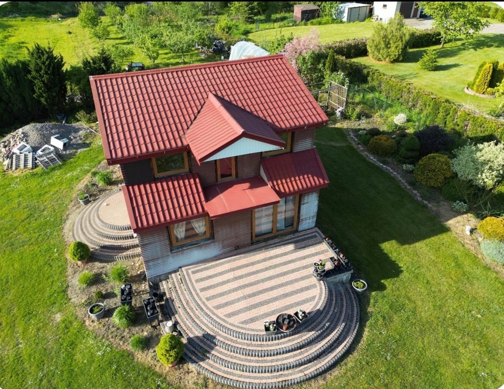 Dom z czerwonym dachem widziany z lotu ptaka, z tarasem wyłożonym kostką w kształcie wachlarza i zadbanym ogrodem.