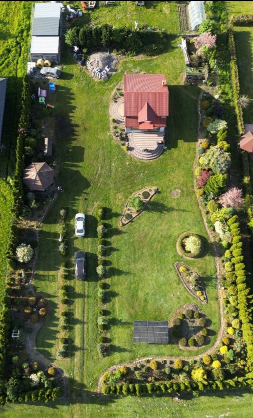 Widok z góry na zadbany ogród z domem z czerwoną dachówką, dwoma samochodami na podjeździe, równo przyciętymi żywopłotami i ozdobnymi rabatami kwiatowymi.