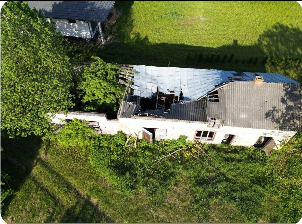 Zdjęcie z lotu ptaka przedstawiające zniszczony, opuszczony budynek z dziurawym dachem i bujną roślinnością wokół.
