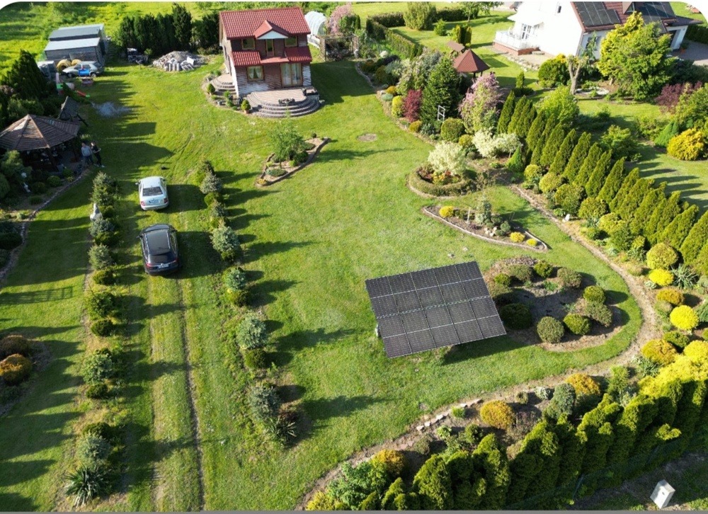 Widok z lotu ptaka na duży ogród z panelem słonecznym, dwoma samochodami na podjeździe i domami w tle.