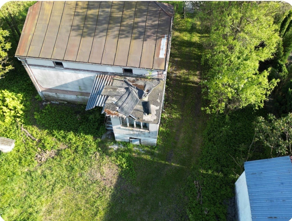 Zdjęcie z lotu ptaka przedstawia zaniedbany budynek z zardzewiałym dachem, częściowo zniszczonymi fragmentami pokrycia i porośniętą roślinnością wokół.