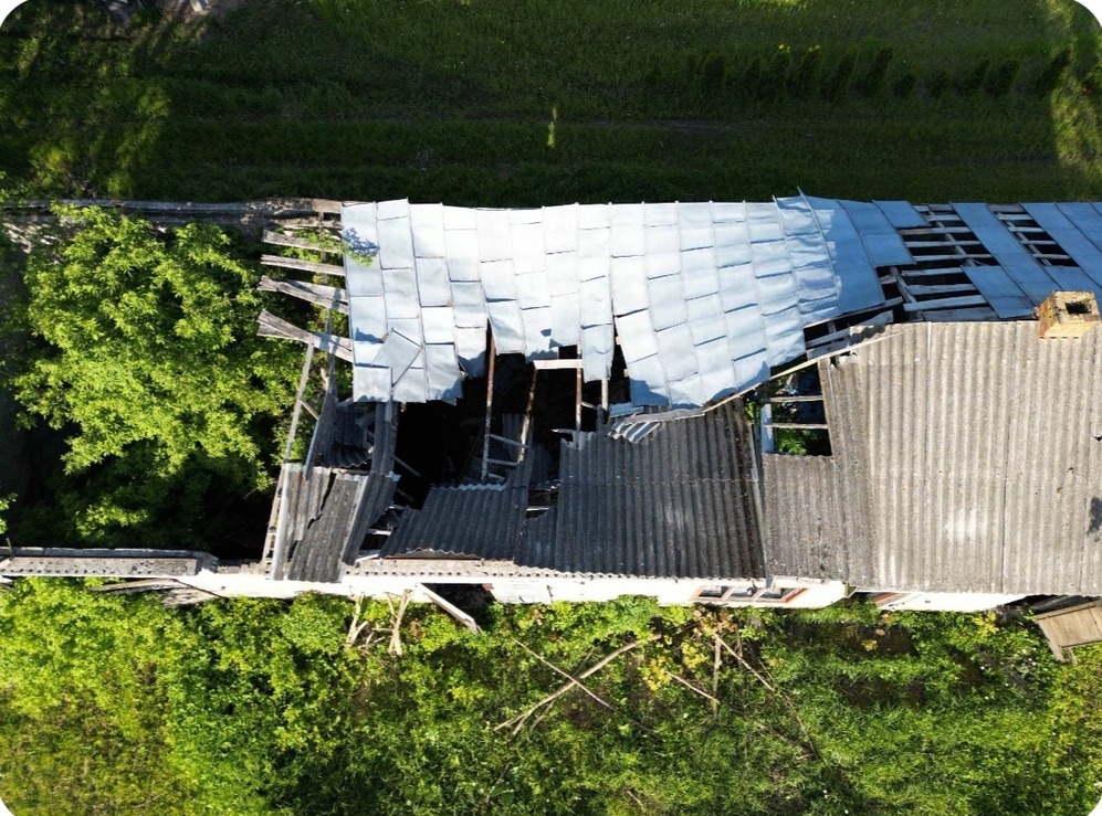 Zdjęcie z drona przedstawia zniszczony budynek z dziurawym dachem, otoczony zielenią, ukazujący stan zaniedbania i potrzebę remontu.