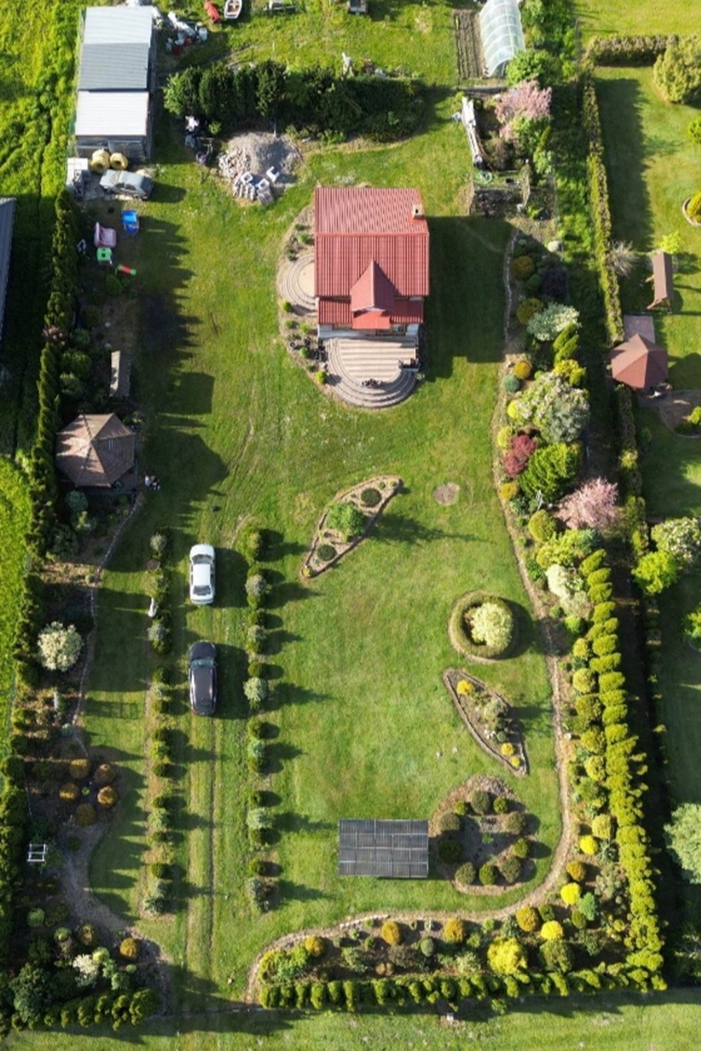 Zdjęcie z drona przedstawiające zadbany ogród z domem, dwoma samochodami, panelami słonecznymi i altaną, otoczony żywopłotem.