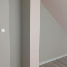 adamczakrenovaties - Pomalowane na szaro ściany z białymi listwami przypodłogowymi i drewnopodobną podłogą. Fragment różowego filaru. Widoczny włącznik światła.