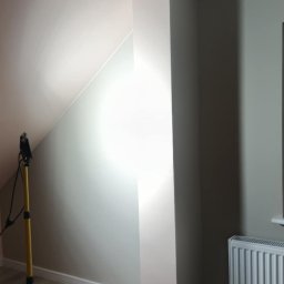 adamczakrenovaties - Świeżo pomalowane wnętrze w Sulmierzycach z lampą roboczą. Widoczne ściany w odcieniach szarości i beżu, podłoga imitująca drewno oraz grzejnik. Gotowe do dalszych prac.