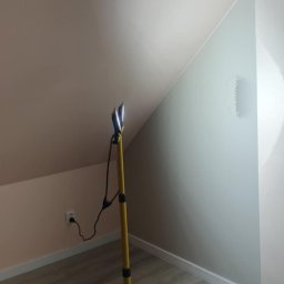 adamczakrenovaties - Pokój na poddaszu po malowaniu ścian na dwa kolory, oświetlony lampą na statywie. Widoczne skośne ściany i podłoga imitująca drewno.