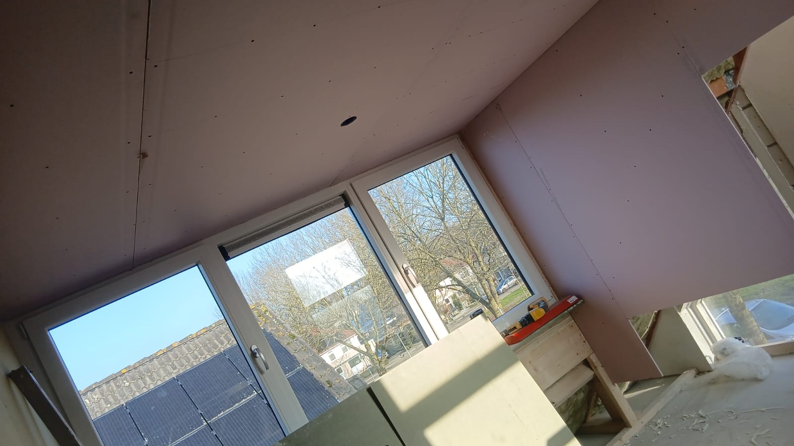 Montaż płyt gipsowo-kartonowych na suficie i ścianach poddasza, widok na okno z drzewami. Widoczne narzędzia i fragmenty konstrukcji.