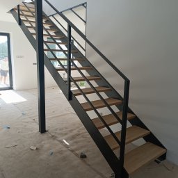 Metal Boys - Nowoczesne schody z czarną metalową konstrukcją i drewnianymi stopniami, widok z dołu, zabezpieczona podłoga w trakcie montażu.