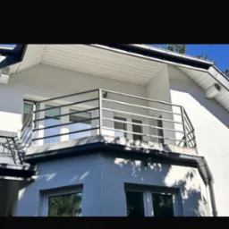 Biały budynek z balkonem zabezpieczonym prostą, metalową balustradą z poziomych prętów, widoczne okna i cień drzewa.