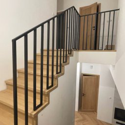 Metal Boys - Nowoczesne schody drewniane z czarną, metalową balustradą o minimalistycznym designie. Widok z dołu, ukazujący konstrukcję i wykończenie. Jasne drewno kontrastuje z czernią.