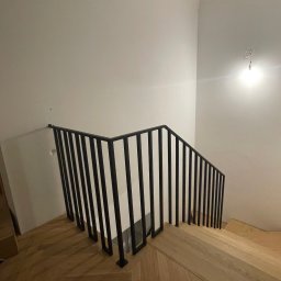 Metal Boys - Minimalistyczna, czarna balustrada metalowa z pionowymi szczeblami, zamontowana na drewnianych schodach w jasnym wnętrzu. Ujęcie z góry, widoczny fragment parkietu.