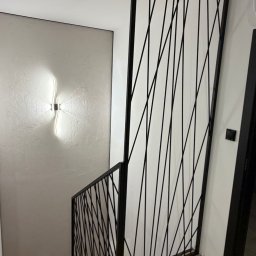 Metal Boys - Nowoczesna klatka schodowa z drewnianymi stopniami i czarną, metalową balustradą o unikalnym, geometrycznym wzorze. Na teksturowanej ścianie świeci designerska lampa.
