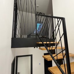 Metal Boys - Nowoczesne schody wewnętrzne z jasnymi, drewnianymi stopniami i czarną metalową balustradą o geometrycznym wzorze z ukośnych prętów. Widoczna pasująca barierka na antresoli.