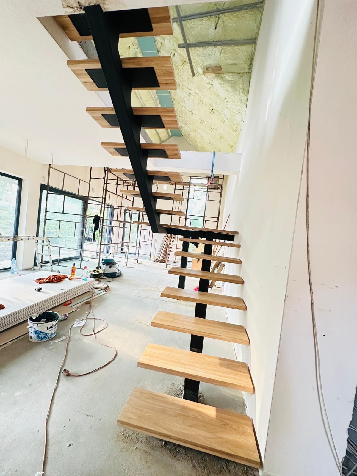 Wnętrze w trakcie wykończenia z drewnianymi schodami na metalowej konstrukcji. Widoczne narzędzia i materiały budowlane. Surowy stan ścian i sufitu.