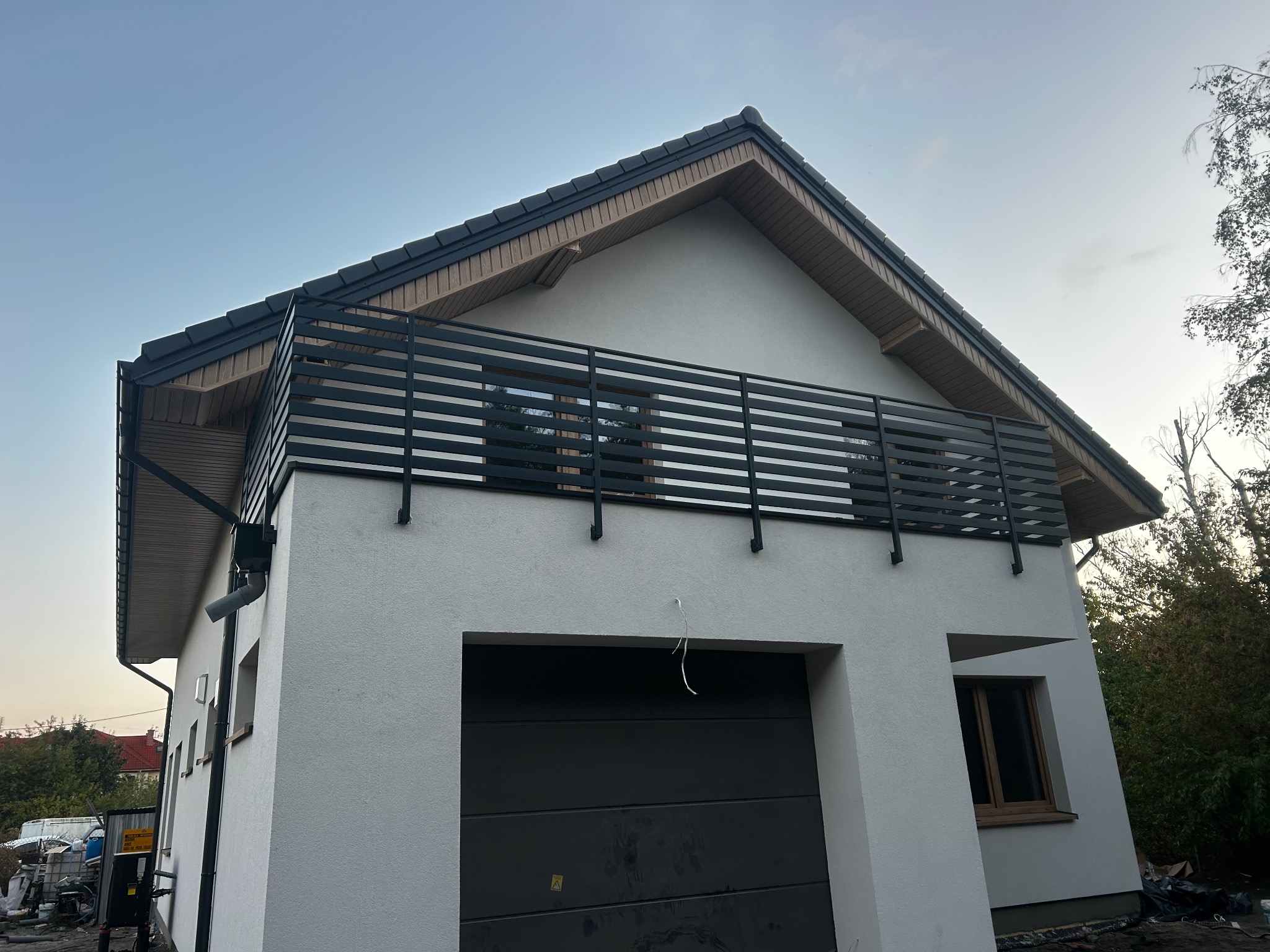 Nowoczesny dom z balkonem zabezpieczonym czarną, poziomą balustradą metalową. Minimalistyczny design, jasna elewacja, ciemna brama garażowa.