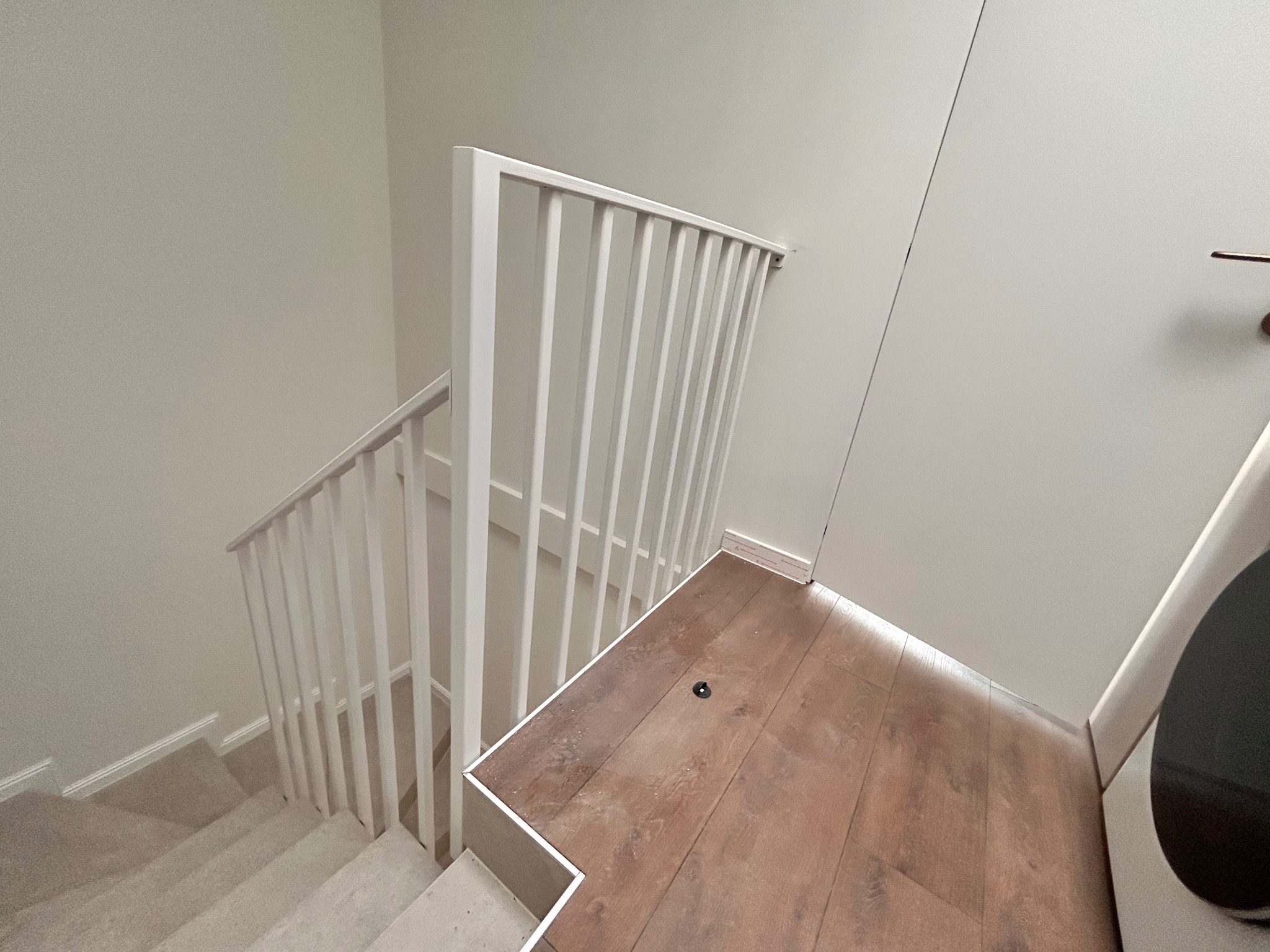 Białe, minimalistyczne schody z balustradą z pionowych szczebli, widziane z góry. Jasne wnętrze, drewniana podłoga na podeście. Ujęcie podkreśla prostotę i nowoczesny design.