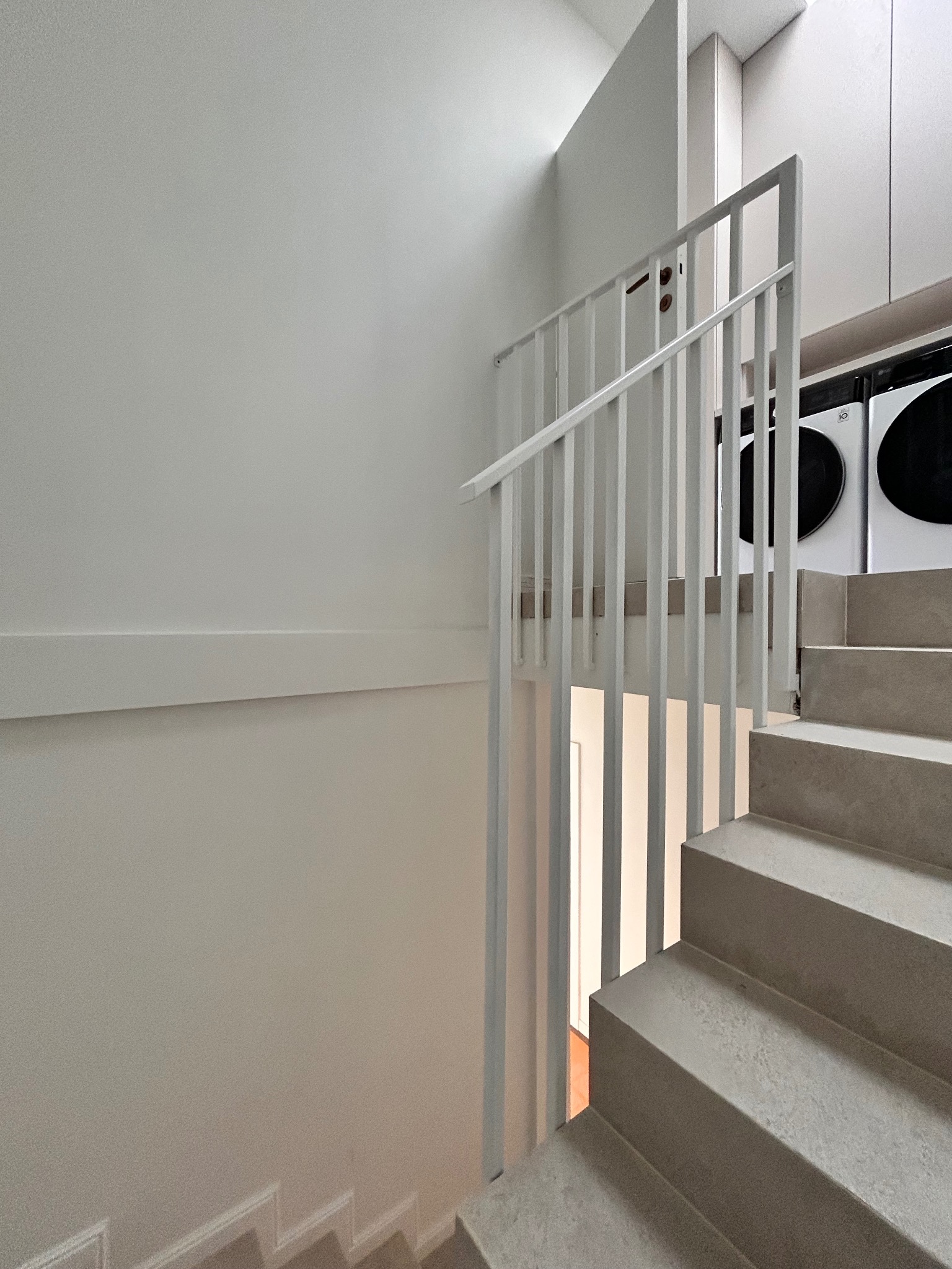 Minimalistyczne schody z jasnoszarego kamienia z białą, metalową balustradą. W tle widoczne pralki, co sugeruje umiejscowienie w pralni lub pomieszczeniu gospodarczym.