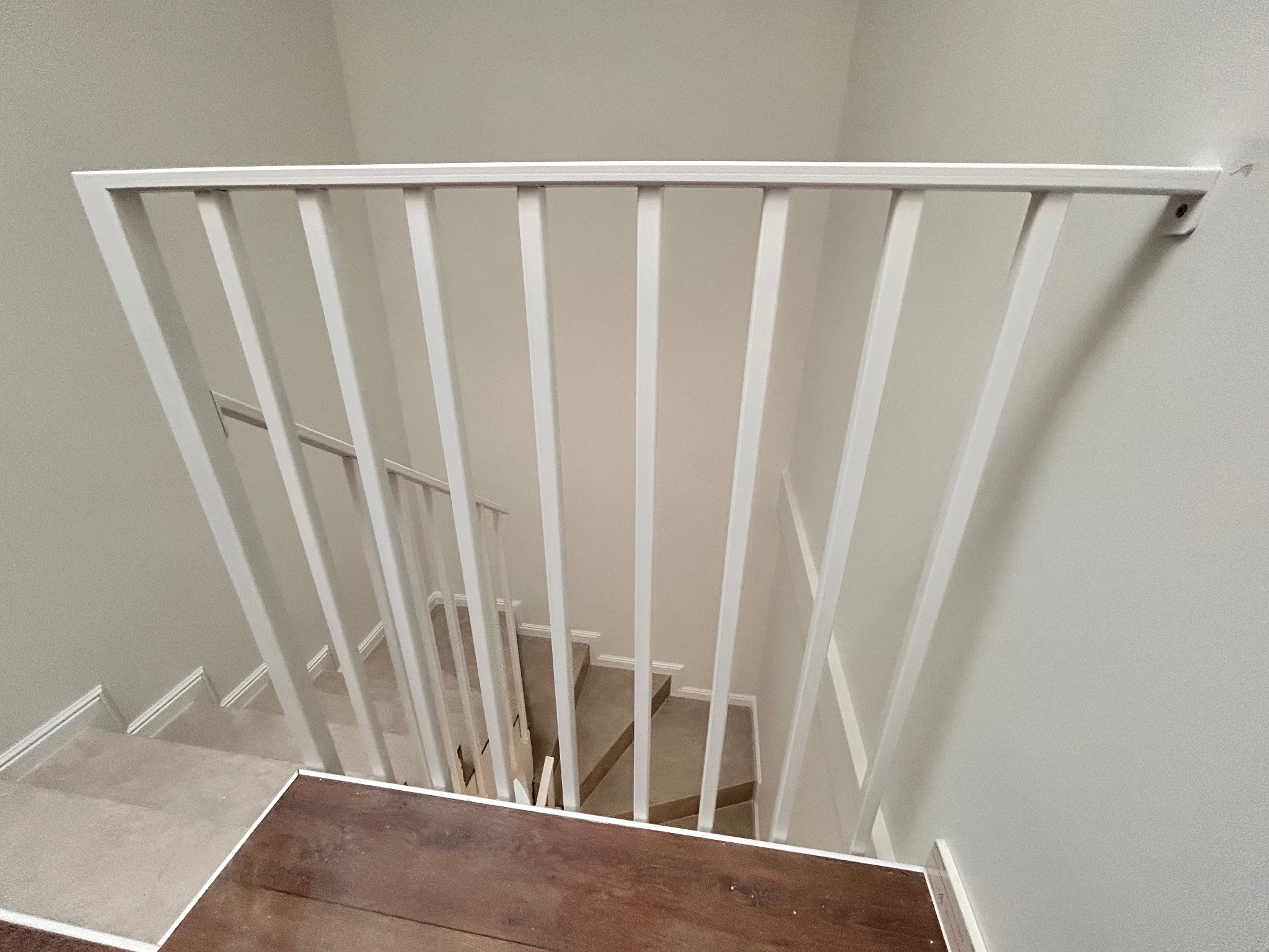 Biała, metalowa balustrada schodowa z pionowymi szczeblami, widok z góry na drewniany podest i schody wyłożone jasną wykładziną. Minimalistyczny design.