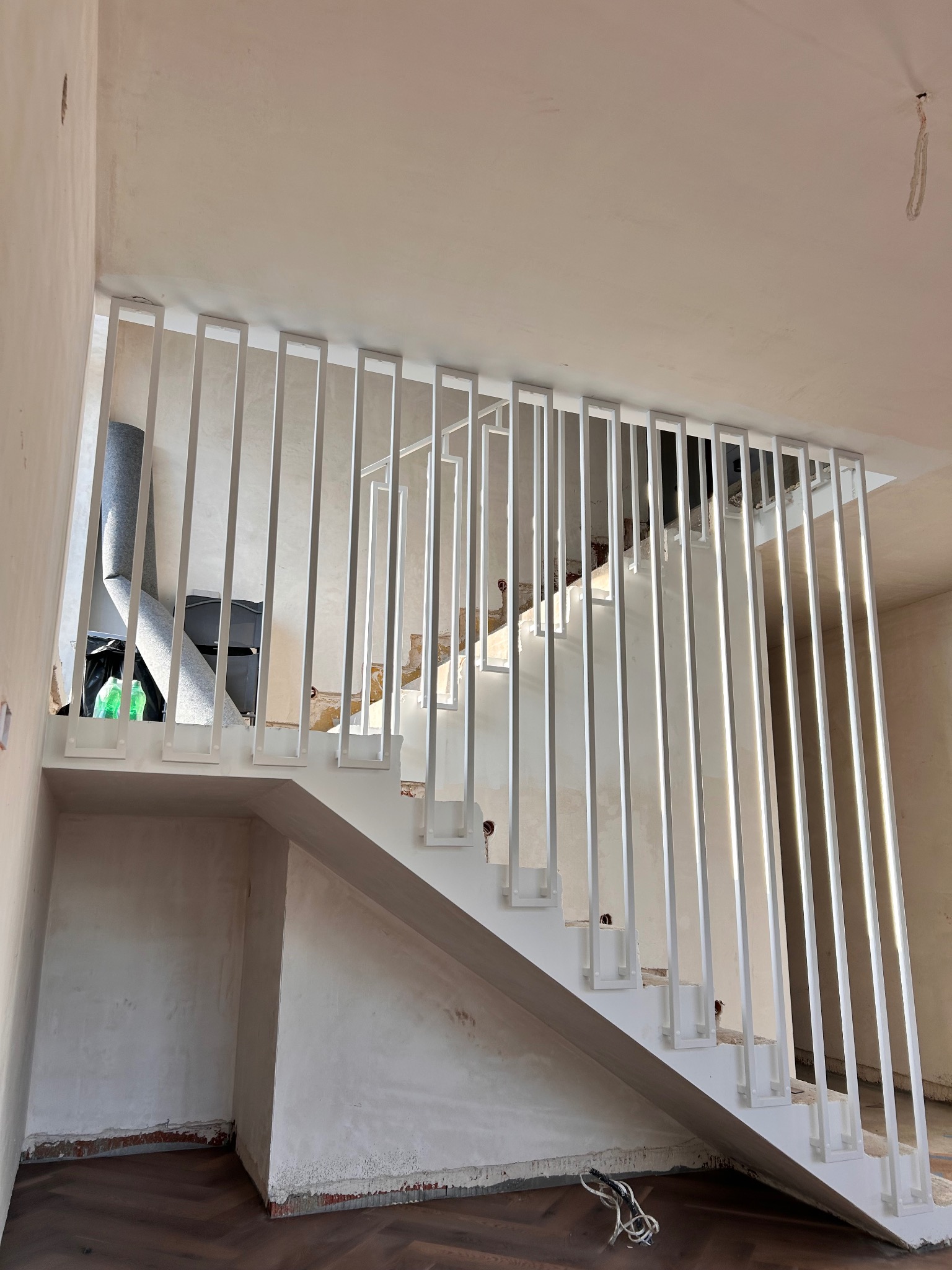 Białe schody z nowoczesną, minimalistyczną balustradą z pionowych, prostokątnych elementów. Widoczne niedokończone ściany i podłoga zabezpieczona folią.