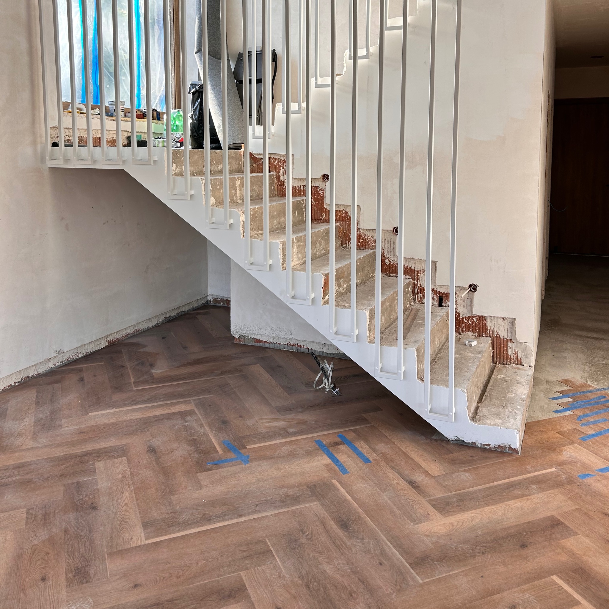 Białe schody z metalową balustradą w trakcie remontu. Widoczne uszkodzenia ścian i podłoga ułożona w jodełkę. Fragment okna z niebieską folią w tle.