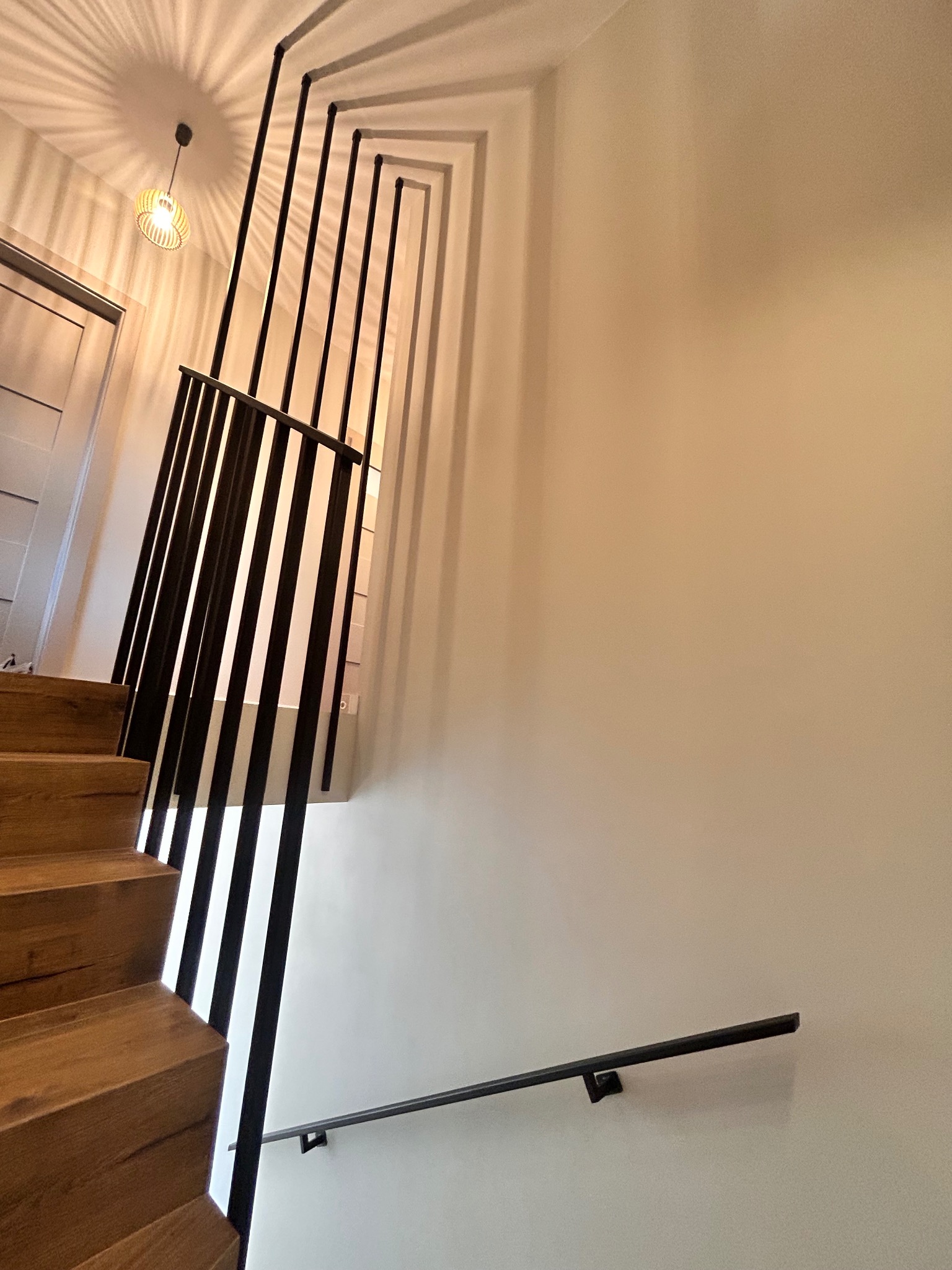 Nowoczesne schody drewniane z czarną, minimalistyczną balustradą metalową. Ujęcie z dołu, widoczne oświetlenie sufitowe i fragment ściany.